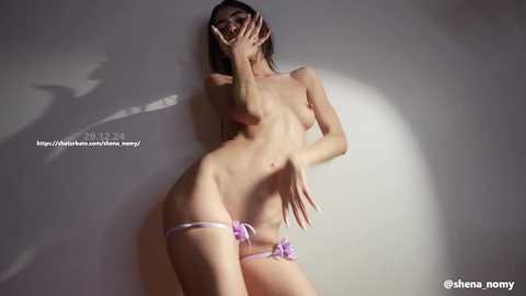 Video of a slender, topless Asian woman with small breasts and fair skin, wearing a pink thong, standing against a white wall, fingers covering her mouth.