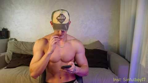 Media: Video of a shirtless, muscular man with a light skin tone wearing a baseball cap and holding a smartphone, standing in a modern living room with beige couches and sheer curtains.