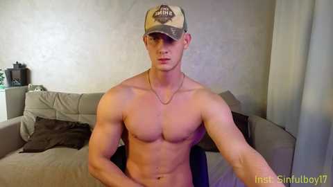 Media: Video of a shirtless, muscular young white man with fair skin, wearing a camo hat, sitting on a beige couch with a brown pillow. The background features a textured wall and a black lamp on a table.