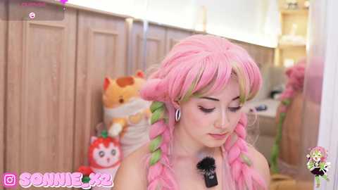 Media: Video of a young woman with pink hair in braids, wearing a pink dress, standing in a cozy room with wooden cabinets and plush toys.