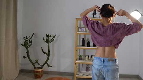 Media: Video of a woman with light brown skin, in a casual setting, tying her hair up. She wears a cropped purple sweater and blue jeans. Behind her is a wooden shelf with plants and bottles, and a cactus in a pot.