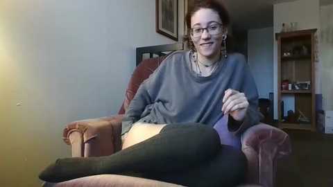 Media: A young woman with glasses, short brown hair, and fair skin, wearing a gray sweater and black leggings, sits comfortably on a brown armchair in a cozy living room.