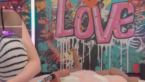 Media: Video of a person holding a black and white striped hat, with a colorful, graffiti-style \"LOVE\" mural in the background, featuring splattered paint.