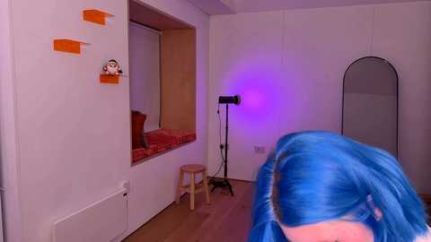 Media: Video of a white-walled room with a blue-haired woman's back, wooden stool, orange shelves, and purple light; toy figures and a mirror in the background.