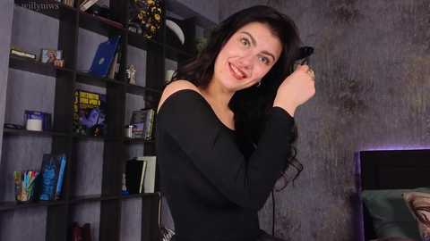 Media: Video of a smiling woman with long black hair, wearing a black off-shoulder top, fixing her hair in a modern, dimly-lit room with bookshelves and a TV in the background.