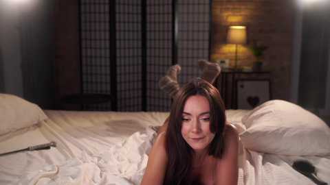 Media: A video of a woman with long brown hair lying on a bed, wearing antler headgear, surrounded by white pillows and sheets. The background features a dimly lit room with a black folding screen and a table lamp.