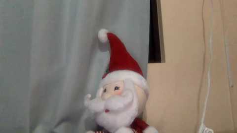 Media: A video of a plush Santa Claus doll with a red hat and white fur trim, placed against a light blue curtain. The background includes a white wall with a small window and a visible electrical cord.