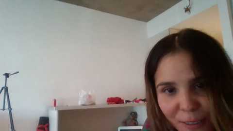 Media: A candid video of a young woman with light brown hair and light skin, smiling slightly, in a minimalist room with white walls, a tripod, and a small shelf with red items and a tissue box.