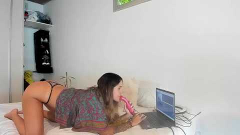 Media: Video of a woman with long brown hair, wearing a colorful blouse and black thong, kneeling on a bed, licking a pink dildo, laptop open in front of her.