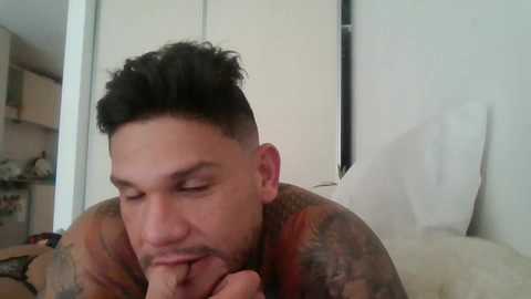 Media: A close-up video of a tattooed, light-skinned man with curly hair, wearing a colorful shirt, in a bedroom with white walls and a bed.