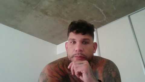 Media: Video of a man with short, curly dark hair, a beard, and tattoos on his arms. He's indoors, against a plain white wall and concrete ceiling, looking contemplative with his hand to his chin.