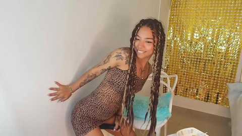 Media: Video of a smiling woman with dreadlocks and tattoos in a leopard print dress, leaning against a white wall with gold tinsel and a blue cushion.