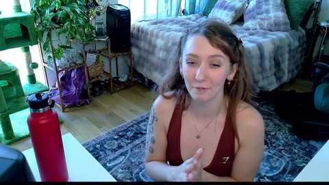 Media: Video of a young woman with light skin, brown hair, and a tattoo, wearing a brown top, sitting on a blue-patterned rug, in a cozy room with a bed, plants, and a red thermos.