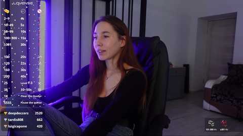 Media: A video of a young, fair-skinned woman with long red hair, seated in a black leather chair, wearing a black top, in a dimly lit room, with a live stream overlay displaying her follower count and chat.