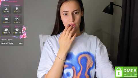 Media: Video of a young woman with long dark hair, light skin, wearing a blue SpongeBob SquarePants t-shirt, biting her finger, in a dimly lit room with a gray wall, black lamp, and a digital clock showing 2:05.