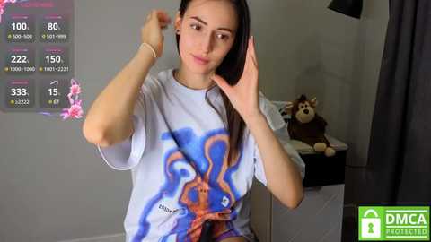 Media: Video of a young woman with long dark hair, wearing a blue and orange graphic tee, adjusting her headphones in a minimalist bedroom.