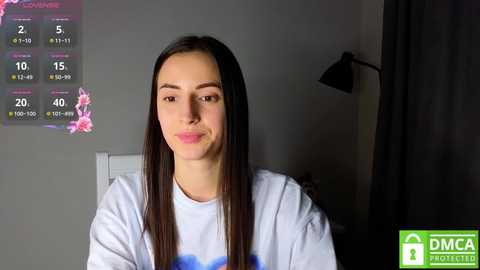 Media: Video of a smiling young woman with straight, long dark hair, wearing a light blue t-shirt, indoors with a calendar showing 2/5 and 2/10.