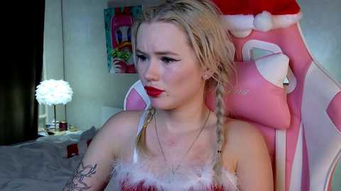 Media: Video of a fair-skinned woman with blonde braided hair in a pink gaming chair, wearing a white feathered top, with a red and white Santa hat, looking pensive. Background includes a colorful wall art and a white lamp.