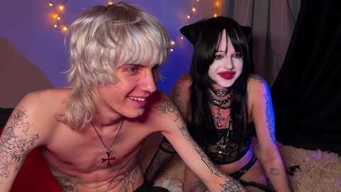 Media: Video of a topless man with platinum blonde hair, tattoos, and a crucifix necklace, and a woman with black hair, cat ears, and a black choker, in a dimly lit room with fairy lights.