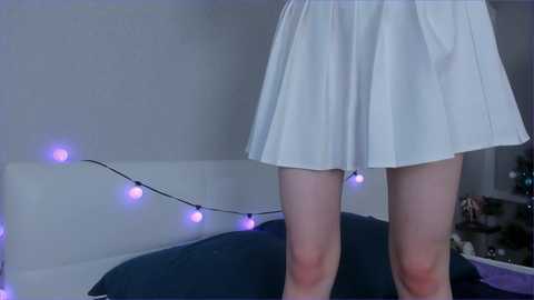 Media: Video of a woman wearing a white pleated skirt, standing with legs apart, in a dimly lit room with purple string lights, white walls, and a bed with dark bedding.