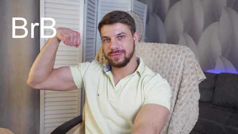 Media: Video of a bearded, muscular man with short brown hair flexing his bicep, wearing a yellow polo shirt. Background features a grey couch, beige wall, and white shuttered window. \"BrB\" text in the top left corner.