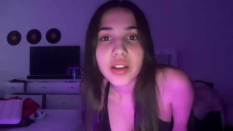 Media: A video of a young woman with long, straight dark hair, wearing a black top, in a dimly lit room with purple lighting. She has a neutral expression and is positioned centrally.