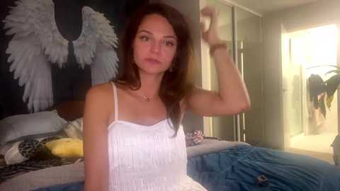 Media: Video of a woman with long brown hair, wearing a white fringed tank top, standing in a bright, cluttered bedroom with a bed, potted plant, and angel wall art.