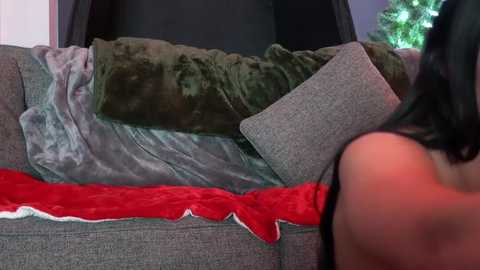 Media: Video of a woman with long black hair, wearing a black top, sitting on a gray sofa. The sofa is covered with a green blanket and a red, fluffy throw with white trim.