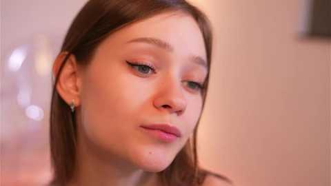 Video of a young woman with fair skin, straight brown hair, and light makeup. She has a neutral expression, wearing a simple top. Background is blurred with soft, warm lighting.