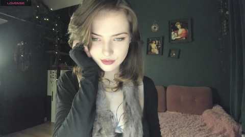 Media: Video of a young woman with light skin, light brown hair, and red lipstick, wearing black gloves, standing in a dimly lit room with a pink bed and framed photos on the wall.