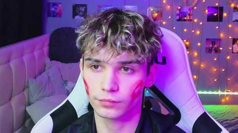 Media: Video of a young man with short, curly, light brown hair, wearing a black and white gaming chair, with purple and orange lights in the background, and photos on the wall.