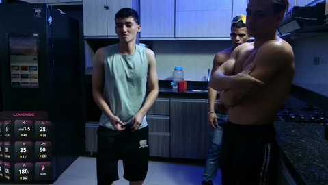 Media: Video of a muscular shirtless man with a tan, flexing in a dimly lit kitchen. Two shirtless men, one with a tattoo, watch. Modern kitchen with white cabinets, black counter, and appliances.