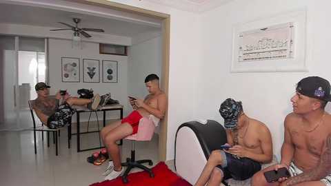 Media: Video: Three shirtless, tattooed men in shorts, wearing black masks, sit on a red couch. One man films with a camera, another plays a video game. Background shows a minimalist, white room with framed art and a ceiling fan.