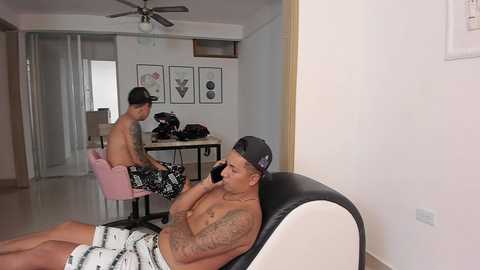 Media: Video of a muscular, tattooed man in a black baseball cap and patterned shorts, seated on a black chair, talking on a phone, in a minimalist, modern living room with white walls, glass sliding doors, and abstract art.