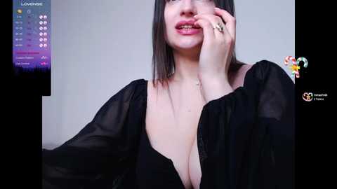 Media: A video of a fair-skinned woman with shoulder-length black hair, wearing a sheer black blouse that reveals her cleavage. She has a ring on her finger, and her hand covers her mouth.