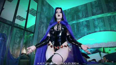 Media: Video of a pale-skinned woman in a glossy black latex bodysuit, red gemstone belt, and purple hooded cape, posing in a dimly lit, ornate room with green lighting.