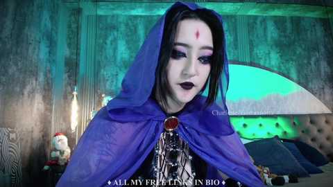 Media: Video of a young woman with pale skin, dark hair, and gothic makeup in a blue hooded cape, set in a dimly lit room with green lighting, and a \"Call My Name\" watermark.