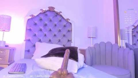 Media: Video of a man's erect penis, uncircumcised, positioned on a bed with white pillows and a gray tufted headboard. The room is softly lit with purple hues, featuring a modern light fixture and a textured wall.