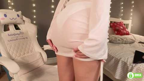 Media: Video of a pregnant woman in a tight white dress, standing in a cozy bedroom with a white gaming chair and a bed with red pillows and a gray blanket.