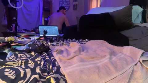 Media: Video of a messy bedroom with a laptop open on a bed, a man sitting shirtless on the bed, a fan, and a blanket with a floral pattern.