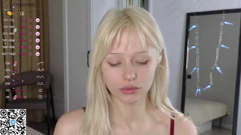 A young, pale-skinned blonde woman with long hair and a nose ring, wearing a red tank top, appears sad in a home setting with a bed and curtains in the background.