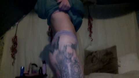 Media: Video of a person lifting their shirt to reveal intricate, large tattoo of a woman on their thigh. Background shows a dimly lit room with hanging dried red peppers and a bed with brown pillows.