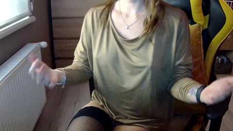 Media: Video of a woman with light skin, wearing a loose beige top, black leggings, and a black wristwatch, sitting on a chair. She has a playful expression, holding a white object in her hands. The background features wooden walls, a radiator, and a yellow backpack.