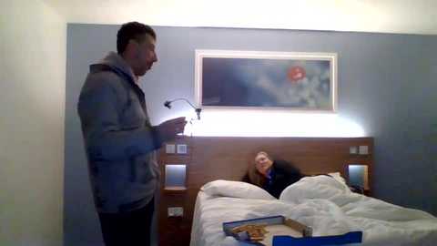 Media: Video of a man in a gray hoodie and woman in black pajamas in a dimly-lit, small hotel room with a wooden headboard, bed, and framed artwork on the wall.