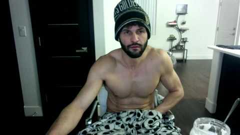 Media: Video of a shirtless, muscular man with a black and white striped beanie, seated on a chair, wearing skull-patterned pajama pants, in a modern, sparsely furnished room with dark wooden floors and white walls.