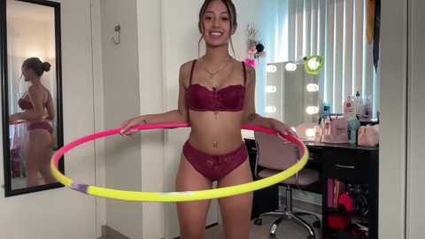 Media: Video of a smiling young woman with medium skin tone and brown hair in burgundy lace lingerie, holding a colorful hula hoop.