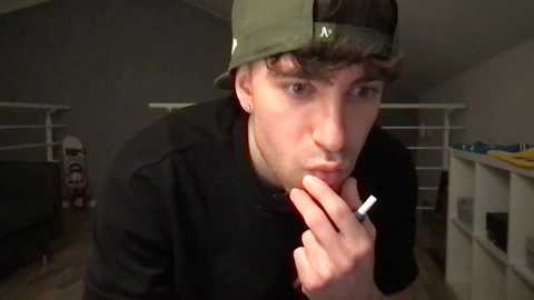 Media: Video of a young man with short brown hair, wearing a green baseball cap and black hoodie, smoking, indoors, dimly lit room with a white shelf and a stuffed toy in the background.