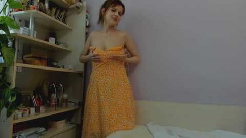 Media: Video of a light-skinned woman with brown hair, wearing a strapless orange dress, adjusting it in a simple bedroom with light-colored walls, a potted plant, and a wooden shelf filled with various items.