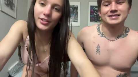 Media: Video of a smiling couple, both shirtless, with tattoos. The woman, with long brown hair, wears a pink bra. The background shows framed pictures.