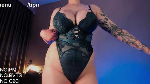 Media: Video of a curvy woman in a dark green lace bodysuit with a plunging neckline, showcasing large breasts and a tattooed arm. Background includes a dimly lit room with a blue light.